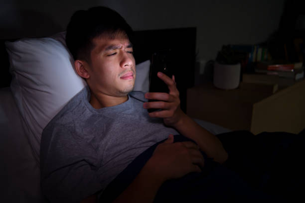 Young man having sore and tired eyes when using a smartphone while lying in bed at night