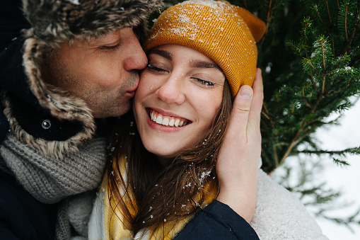 Happy man kissing his smiling woman in a cheek in front of a fir tree in the winter. Close up.