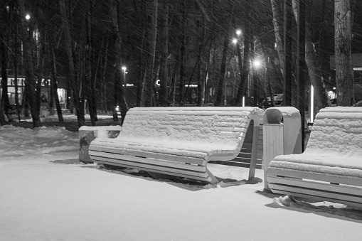 City landscape. Night. Snowfall. Snow-covered benches in a city park, illuminated by a lantern.