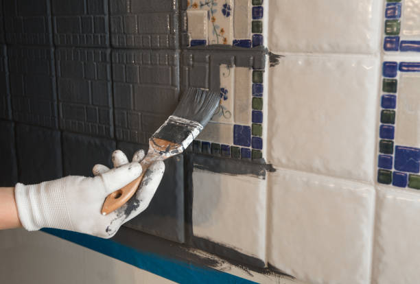 Repainting old dated kitchen ceramic tile back wall with modern gray chalk paint indoors at home. Giving old kitchen new look concept. Hand holding a paint brush tool.