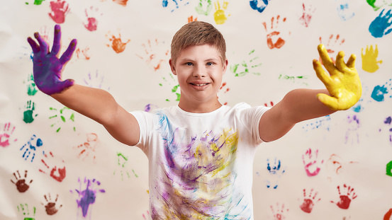 Happy disabled boy with Down syndrome smiling at camera while reaching out his hands painted in colorful paints ready for hand prints on the wall. Children with disabilities and special needs concept