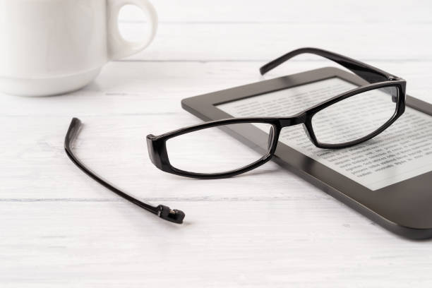 istock Eyeglasses with one temple broken off the rim on a e-reader over white wood desk. Breakage of vision correction glasses for reading, remote learning and computer work. 1292280218