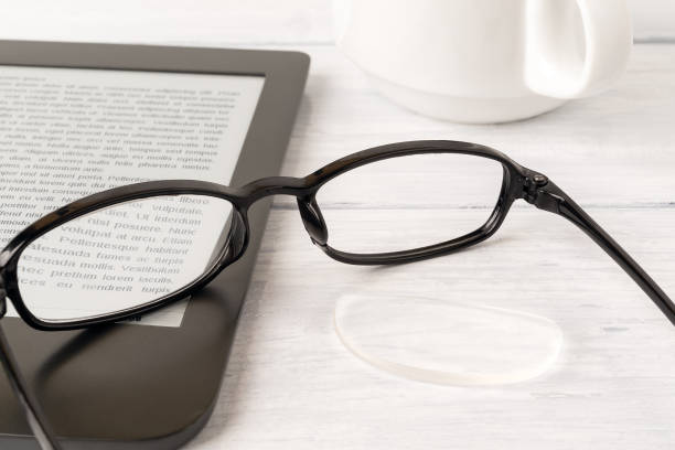 istock Eyeglasses with one lens fell down from the rim on a e-reader over white wood desk. Breakage of vision correction glasses for reading, remote learning and computer work. 1292280226