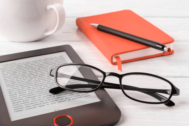 istock Eyeglasses on e-reader, orange note pad and cup on a white wooden desk. Vision correction glasses for reading and computer work. E-reading for pleasure and education. 1292280240