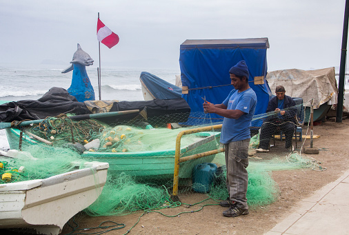 Lima, Peru - June 18, 2015: Commercial fishermen mend their nets by their fishing boats that are docked on the Pacific Ocean beach in Miraflores.