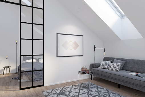 Ático moderno con póster horizontal en una pared blanca entre un sofá gris con mesa de centro y una partición de cristal. Hay una ventana en la azotea y una alfombra en el suelo de madera de la habitación. photo