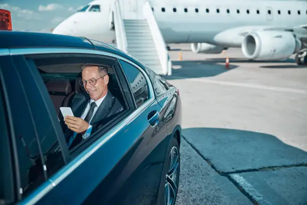 Photo of Cheerful businessman with smartphone in car at airport