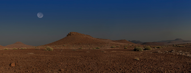 During an early morning drive in Damaraland, Namibia,  the moon is setting over hills above the red rock desert.