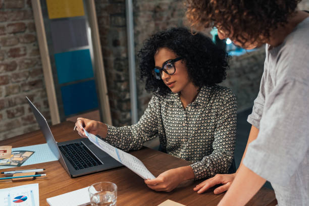 istock Women in Business: Two Young Businesswoman Using Technology and Analyzing Reports in an Office 1292307469