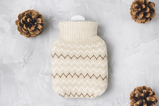 Gray mini silicone hot water bottle and knitted cover for hand or body warming or for winter illness on gray background. Keeping warm in winter outdoor or at home in bed concept