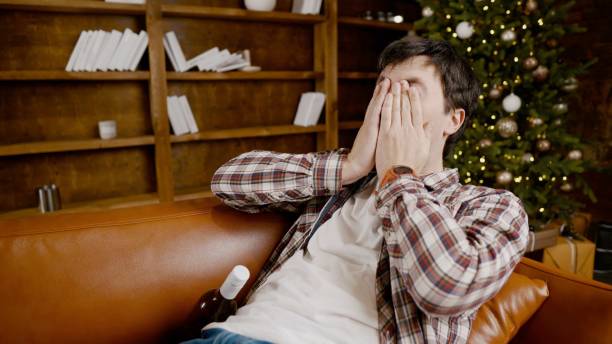 istock Young male alcoholic requires medical and psychological help. Man addicted alcohol and cannot stay on couch at home on Christmas and New Year Eve. Drunk guy drink wine from bottle near Christmas tree 1292314511