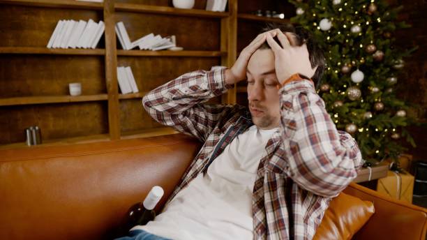 istock Young male alcoholic requires medical and psychological help. Man addicted alcohol and cannot stay on couch at home on Christmas and New Year Eve. Drunk guy drink wine from bottle near Christmas tree 1292314527