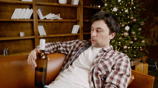 istock Young male alcoholic requires medical and psychological help. Man addicted alcohol and cannot stay on couch at home on Christmas and New Year Eve. Drunk guy drink wine from bottle near Christmas tree 1292314545