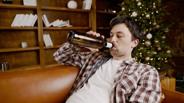 istock Young male alcoholic requires medical and psychological help. Man addicted alcohol and cannot stay on couch at home on Christmas and New Year Eve. Drunk guy drink wine from bottle near Christmas tree 1292314551