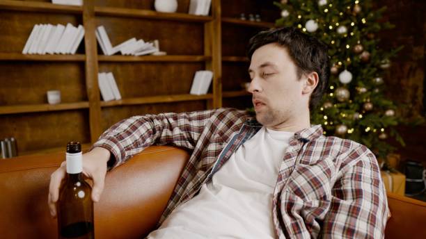 istock Young male alcoholic requires medical and psychological help. Man addicted alcohol and cannot stay on couch at home on Christmas and New Year Eve. Drunk guy drink wine from bottle near Christmas tree 1292314564