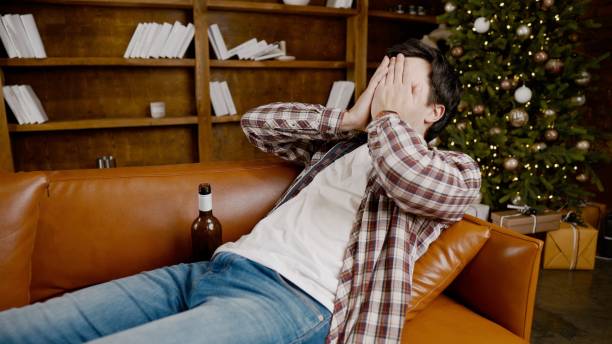 istock Young male alcoholic requires medical and psychological help. Man addicted alcohol and cannot stay on couch at home on Christmas and New Year Eve. Drunk guy drink wine from bottle near Christmas tree 1292315191