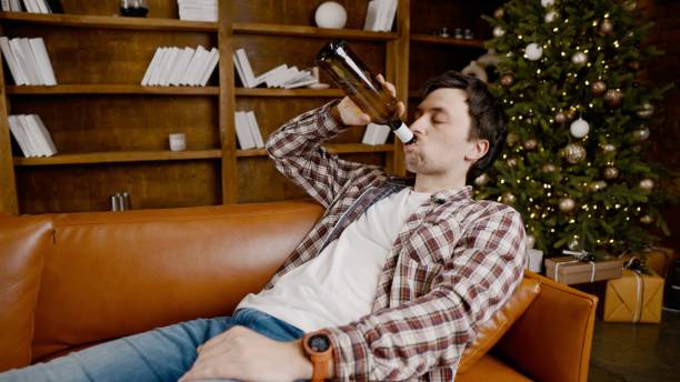 istock Young male alcoholic requires medical and psychological help. Man addicted alcohol and cannot stay on couch at home on Christmas and New Year Eve. Drunk guy drink wine from bottle near Christmas tree 1292315201