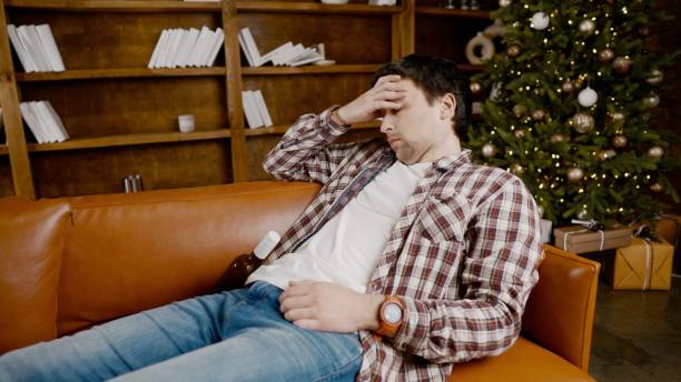 istock Young male alcoholic requires medical and psychological help. Man addicted alcohol and cannot stay on couch at home on Christmas and New Year Eve. Drunk guy drink wine from bottle near Christmas tree 1292315341