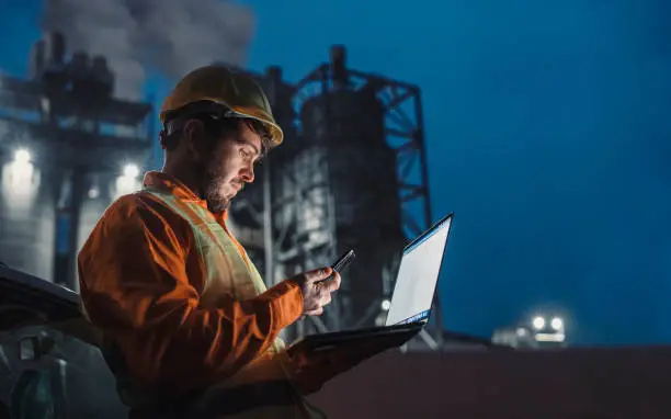 Side view of engineer working nightshift and using technology in front of petroleum industrial factory and using laptop and smart phone.