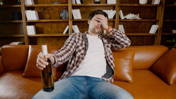 istock Young male alcoholic requires medical and psychological help. Man addicted alcohol and cannot stay on couch at home on Christmas and New Year Eve. Drunk guy drink wine from bottle near Christmas tree 1292315465