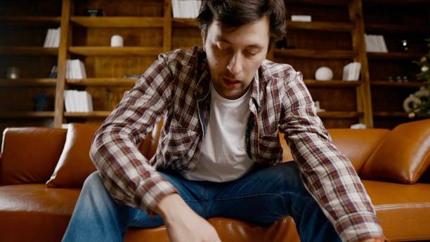 istock Young male alcoholic requires medical and psychological help. Man addicted alcohol and cannot stay on couch at home on Christmas and New Year Eve. Drunk guy drink wine from bottle near Christmas tree 1292316017