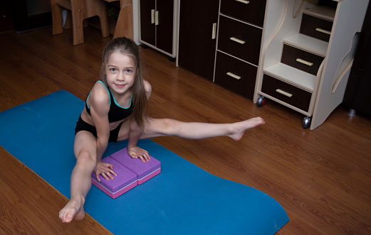 girl 8 years old with long hair in black sportwears do exercises around home with two blocks, stretching at home without trainer
