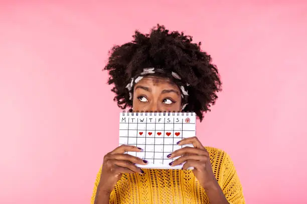 Photo of Portrait of a funny young African American girl in hiding behind a menstrual periods calendar and looking away at copy space isolated over pink background. Female Period calendar
