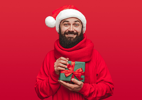 Glad bearded guy in Santa hat demonstrating present and smiling for camera on Christmas day against red background