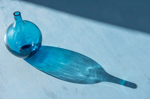 Blue color design glass vase on blue wooden background with sunlight and hard shadow. Large copy space.