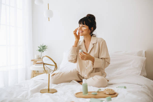 mujer sonriente aplicando crema facial sentada en la cama - cuidado de la piel fotos fotografías e imágenes de stock