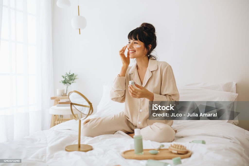 Smiling woman applying face cream sitting on bed Happy woman doing routine skin care at home with beauty products. Woman sitting on bed at home and applying face cream. Skin Care Stock Photo
