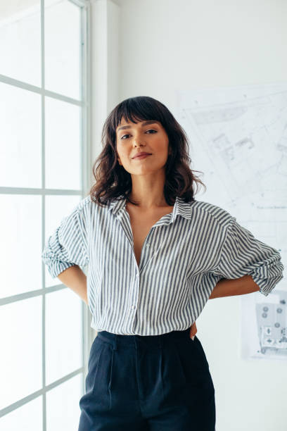 Confident businesswoman standing in office Portrait of businesswoman standing beside the window in office. Female architect standing in office with architecture drawings in the background. architect photos stock pictures, royalty-free photos & images