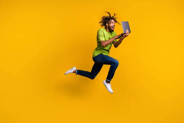 Full length photo portrait of excited programmer with dreadlocks jumping up holding laptop in hands isolated on vivid yellow colored background.
