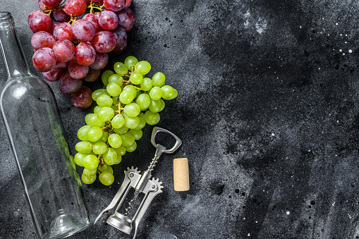 A branch of green and red grapes, an empty bottle, a corkscrew and a cork. Concept of home winemaking. Black background. Top view. Copy space.