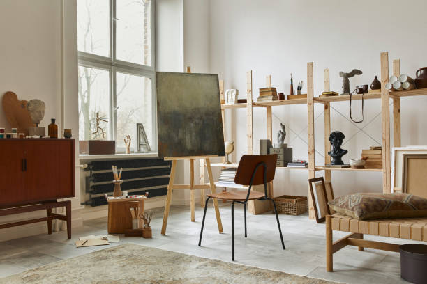 istock Unique artist workspace interior with stylish teak commode, wooden easel, bookcase, artworks, painting accessories, decoration and elegant personal stuff. Modern work room for artist. Template. 1292378135
