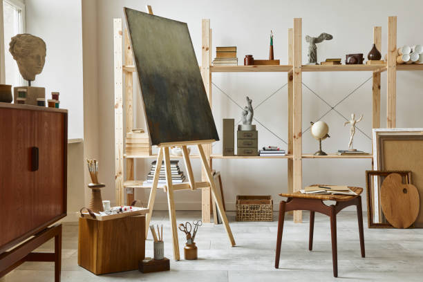 istock Unique artist workspace interior with stylish teak commode, wooden easel, bookcase, artworks, painting accessories, decoration and elegant personal stuff. Modern work room for artist. Template. 1292378149