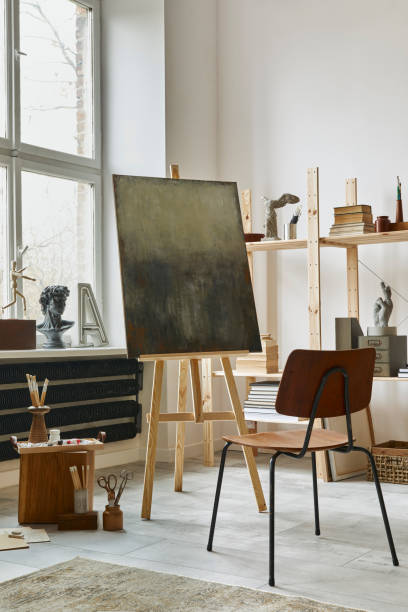 istock Unique artist workspace interior with stylish teak commode, wooden easel, bookcase, artworks, painting accessories, decoration and elegant personal stuff. Modern work room for artist. Template. 1292378666