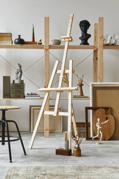 istock Unique artist workspace interior with stylish teak commode, wooden easel, bookcase, artworks, painting accessories, decoration and elegant personal stuff. Modern work room for artist. Template. 1292378674