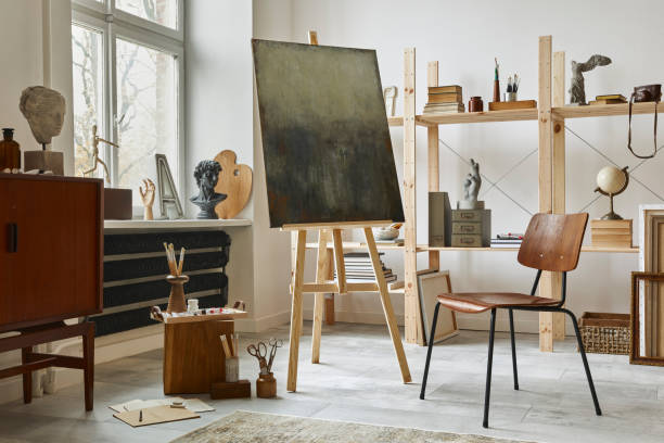 istock Unique artist workspace interior with stylish teak commode, wooden easel, bookcase, artworks, painting accessories, decoration and elegant personal stuff. Modern work room for artist. Template. 1292378677