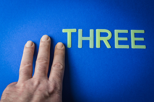 Three human fingers beside the word Three written with plastic letters on blue paper background, concept