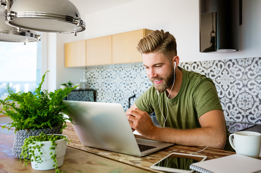 Happy young man wearing green t-shirt and earphone sitting at table in the kitchen and working at home, having video conference.