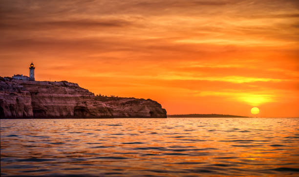 Sun rising over the sea in Algarve, with the rock cliff and caves and the Farol de Anfanzina Lighthouse in Carvoeiro. Portgugal
