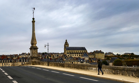 Blois, France, October 28, 2020: A man goes over the Jacques Gabriel Bridge (French: Pont Jacques Gabriel) from 18th century over the Loire River in Blois under cloudy sky. In the background is the Saint-Louis Cathedral. The Loire Valley is listed as UNESCO World Heritage Site.