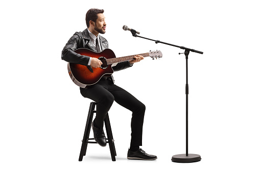 Man in a leather jacket sitting on a chair singing with a microphone on a stand and playing an acoustic guitar isolated on white background