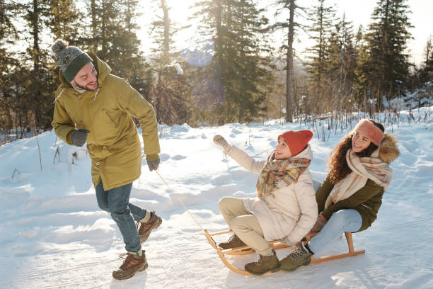 istock Two joyful girls in winterwear sitting on sledge while young man pulling them 1292402234