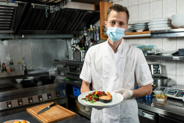 istock Male chef in protective mask holding plate with fried salmon and vegetables 1292402413