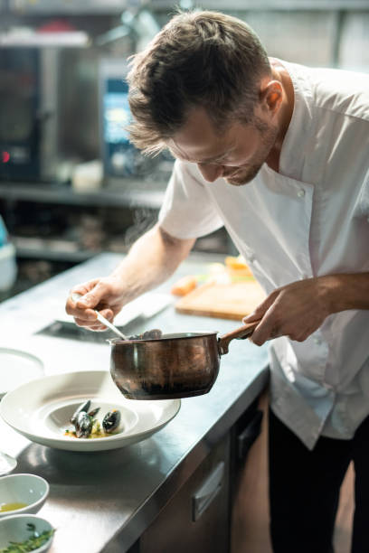 istock Chef in uniform bending by kitchen table and putting boiled mussels on plate 1292402416