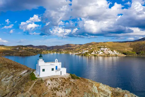 Greece, Kea Tzia island. Aerial drone view of lighthouse and white St. Nicolas church on a rocky cape, cloudy blue sky and transparent rippled sea water background. Summer day, tourist destination.