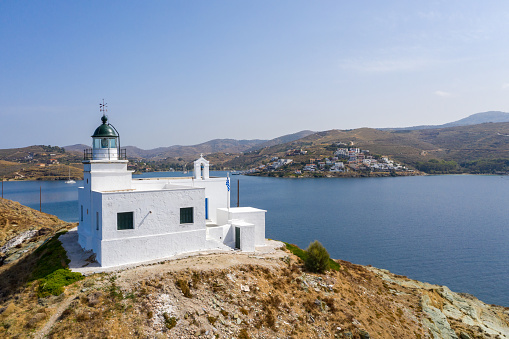 Greece, Kea Tzia island. Aerial drone view of lighthouse and white St. Nicolas church on a rocky cape, clear blue sky and transparent rippled sea water background. Summer, tourist destination.
