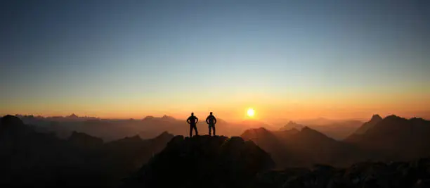 Happy winning success men at sunset or sunrise standing relaxed and are happy for having reached mountain top summit goal during hiking travel trek. Tirol, Austria. Allgau, Bavaria.